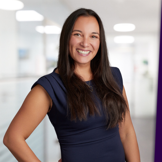 Breit lächelnde Frau mit dunkelbraunen langen Haaren in dunkelblauen eng anliegenden Kleid. Die linkge Hand ist an die Hüften gestützt.