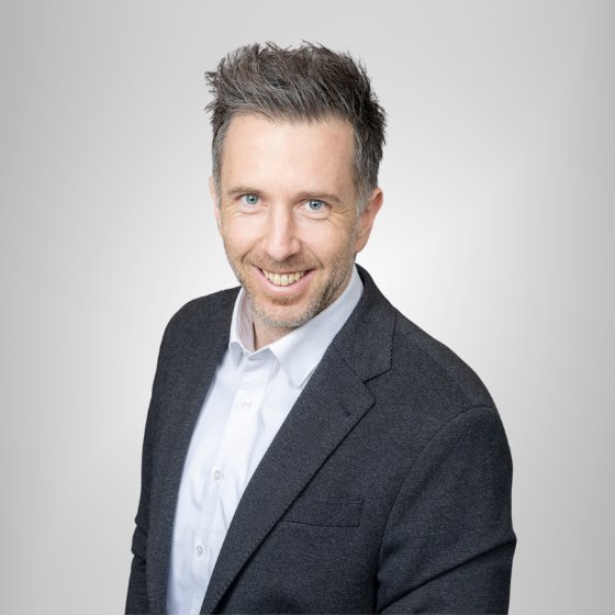 Mann in schwarzem Sakko mit weißem Hemd lächelt in die Kamera. Blick von unten.