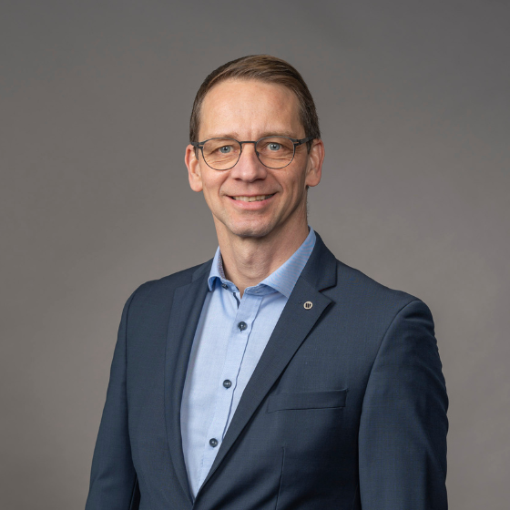 Mann mit Brille lächelt ins Bild. Hellblaues Hemd mit dunkelblauem Blazer. Portraitfoto