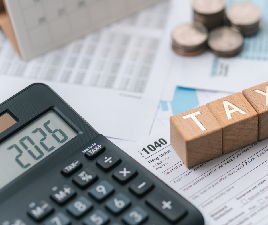 Taschenrechner der die Zahlen 2026 anzeigt liegt auf einem Papierhaufen von wichtigen Unterlagen mit Münzen und Holzbausteinen die "TAX" sagen.