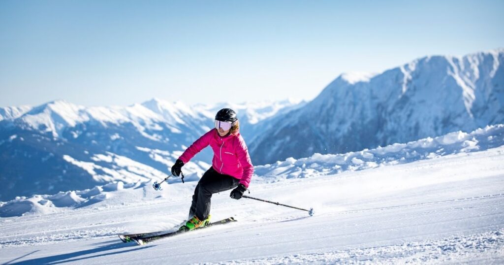 Schneebedeckte Bergkulisse mit Skifahrerin davor die gerade die Piste herunter fährt. Pinke Skijacke, Helm mit Skibrille und neongrüne Skischuhe.
