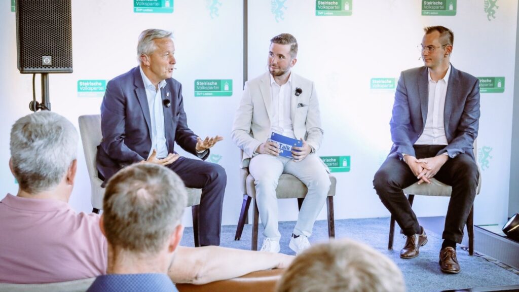 Drei Männer sitzen bei einer Podiumsdiskussion auf einer Bühne. Der Mann links spricht und gestikuliert, der Moderator in der Mitte hält Karten in der Hand, der Mann rechts hört zu. Im Vordergrund ist das Publikum zu sehen, im Hintergrund eine Wand mit Logos der ÖVP Leoben.