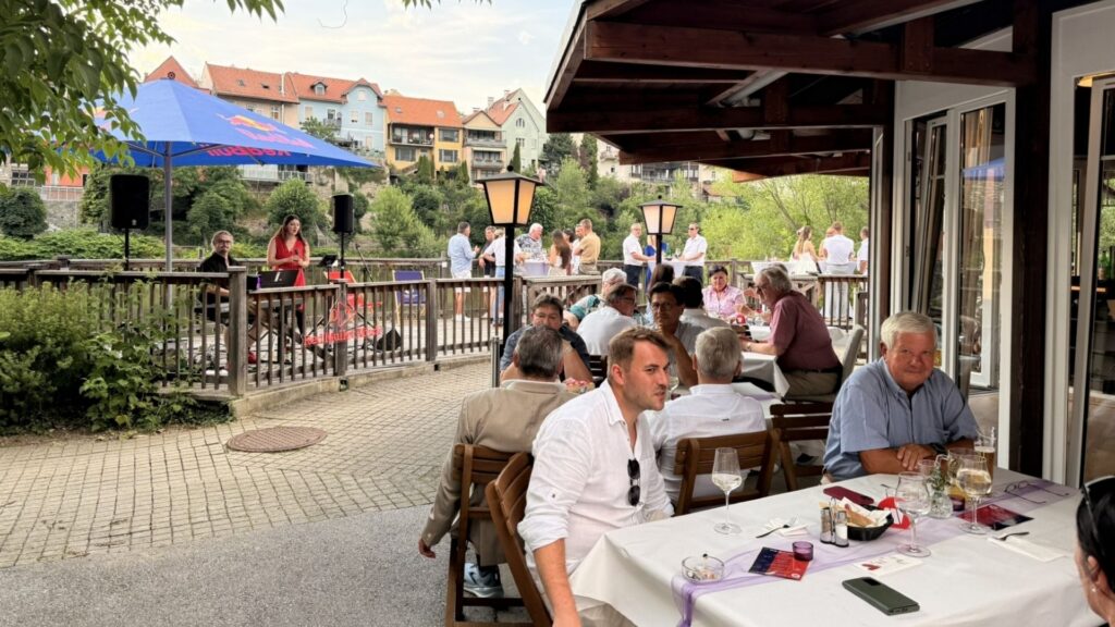 Besucherinnen und Besucher des Sommerfestes des Wirtschaftsbundes Graz-Umgebung sitzen an gedeckten Tischen auf einer überdachten Terrasse und unterhalten sich. Weitere Gäste stehen im Außenbereich am Geländer; im Hintergrund sind Bäume und Häuser zu sehen.